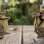A photograph of two ornate, gold-toned 'Golden Bee' dance medals, each with unique dancer figures and bee motifs, resting on a rustic garden table with blue ribbons.